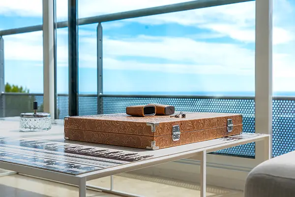 Photo en gros plan d'une mallette en cuir marron posé sur une table en verre dans un salon design et contemporain d'une villa de luxe, avec vue sur la mer par la fenêtre en arrière plan, dans un contexte de photos et vidéos immobilières.